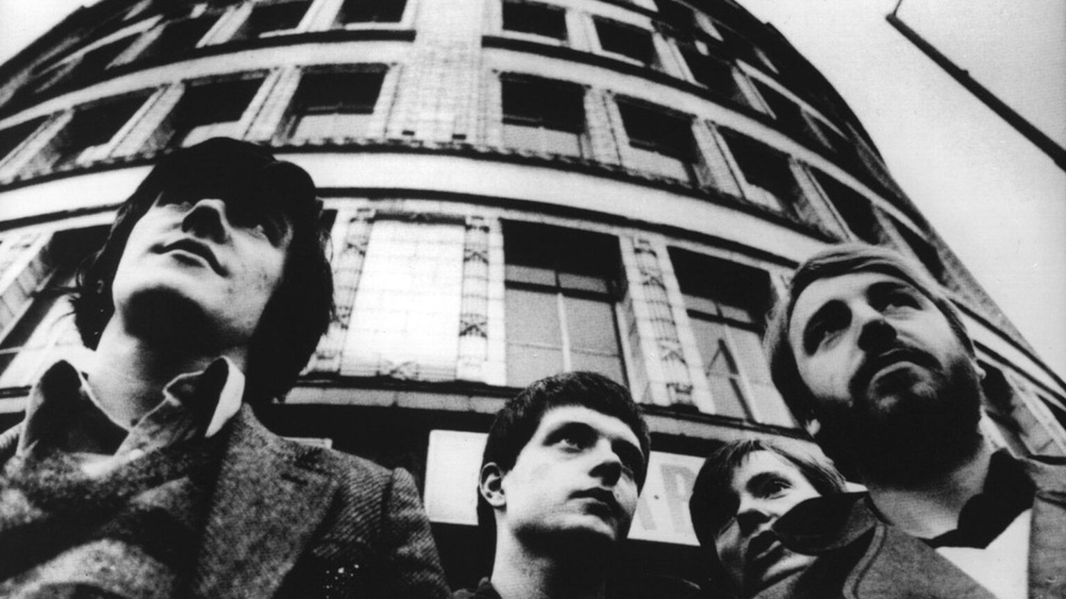 Joy Division: dos álbumes, cuatro años y una influencia desproporcionada Joy Division fotografiados en Manchester: Ian Curtis, Bernard Sumner, Peter Hook y Stephen Morris mirando hacia arriba frente a un edificio curvo.